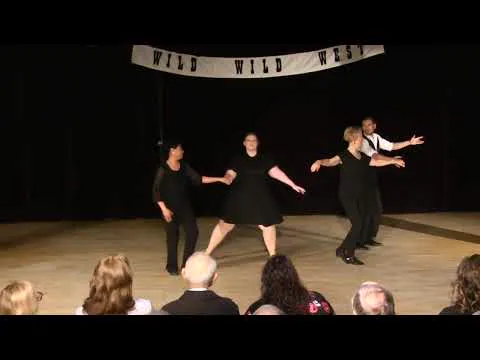 Four dancers in black perform on a stage under a Wild Wild West banner, with the audience in the foreground.