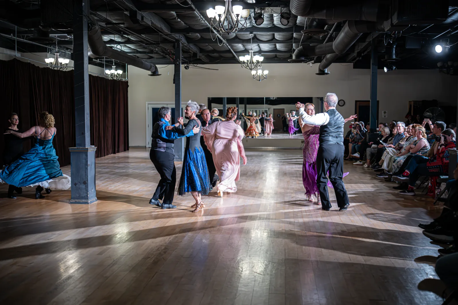 Couples dancing across the Spotlight Ballroom floor under chandeliers, with a mirror wall and audience.