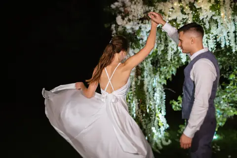 Bride in a flowing white dress twirls under a floral arch at night while the groom watches
