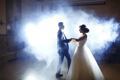 Bride and groom dance hand in hand in glowing fog, lit from behind