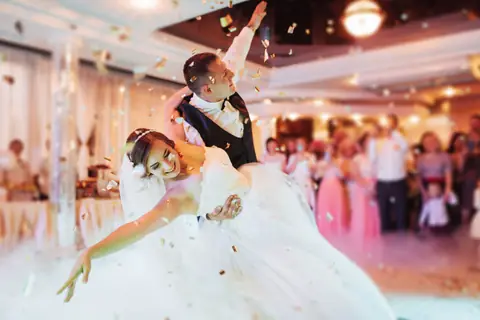Groom dips his bride in a ballroom as confetti falls and guests look on