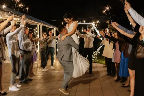 A groom carries his bride between guests holding sparklers at night
