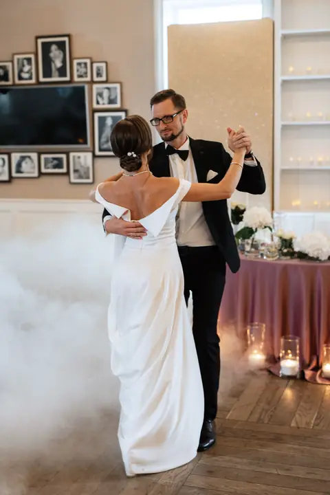 Bride in an off-shoulder gown dances with her groom in a bow tie in a fog-filled room